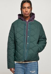 Chaqueta verde acolchada con un patrón curvado, cierre a presión y cuello redondeado, llevada sobre una sudadera con capucha morada.