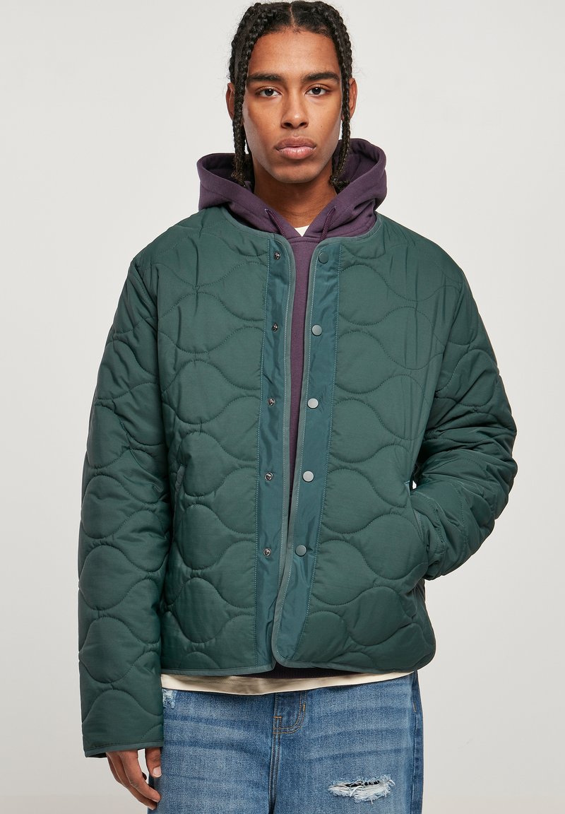 Chaqueta verde acolchada con un patrón curvado, cierre a presión y cuello redondeado, llevada sobre una sudadera con capucha morada.