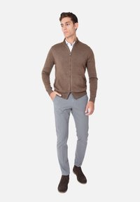 Maglione marrone con zip e lavorazione a maglia, abbinato a chinos grigi chiari e scarpe marroni scure. Il modello si trova su uno sfondo neutro.