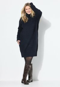 Abito in maglia oversize blu navy con collo alto, abbinato a stivali alti fino al ginocchio in stampa leopardata lucida, indossato sopra collant trasparenti neri.