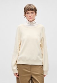 Sudadera color crema con escote redondo y puños acanalados, combinada con un cuello alto ligero. Tejido suave y ajuste cómodo.