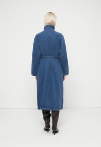 Personne aux cheveux blonds courts portant un long manteau en jean bleu avec ceinture et col montant, associé à des bottes noires montantes à talons, vue de dos.