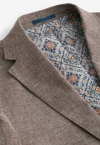 Brauner struktureller Blazer mit einem gemusterten Futter, das blaue, orange und beige Blumenmuster zeigt. Auffällige Details am Kragen und Revers.