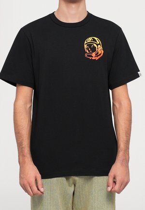 Mann trägt ein schwarzes T-Shirt mit einer kleinen orange-gelben Astronautenhelmgrafik auf der linken Brust und hellen Hosen.