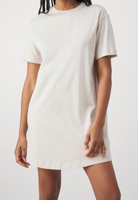Vestido de manga corta, por encima de la rodilla, color beige claro, hecho de algodón con cuello redondo y pequeño logo blanco en el pecho, con una textura suave.