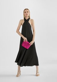 Black halter neck dress with a flowing silhouette. Holds a pink pleated clutch. Worn with strappy, jeweled sandals. Smooth fabric texture.