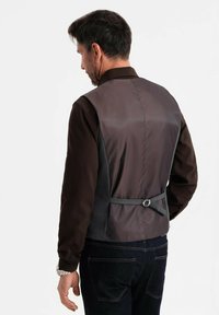 Mann trägt ein dunkles Langarmhemd, eine graue verstellbare Weste und dunkle Jeans, von hinten vor einem schlichten weißen Hintergrund gezeigt.