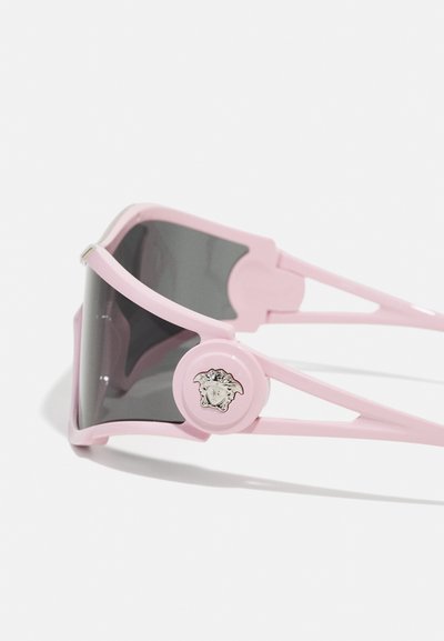 Pink sunglasses with oversized dark lenses, featuring a logo medallion on the frame and angular arms with cut-out detailing.