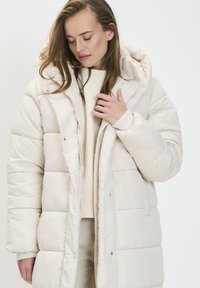 Doudoune beige clair avec capuche, présentant des sections matelassées, des pressions à l'avant et une fermeture éclair. Texture douce, adaptée aux temps froids.