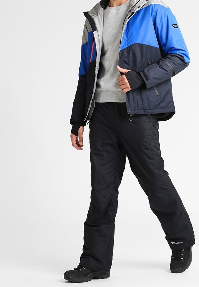 Personne portant une veste bleue, noire et grise, un sweat-shirt gris, un pantalon noir et des chaussures de randonnée noires, debout sur un fond blanc.