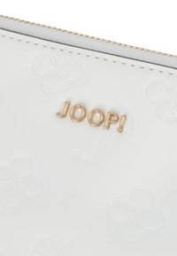 Portefeuille en cuir verni blanc avec un motif floral embossé et un logo "JOOP!" en doré. Fermeture à glissière dorée et doublure en tissu texturé.