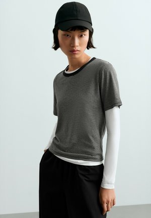 Jeune personne portant une casquette noire, une chemise à manches courtes rayée noir et blanc par-dessus une chemise blanche à manches longues, et un pantalon noir, debout les mains dans les poches.
