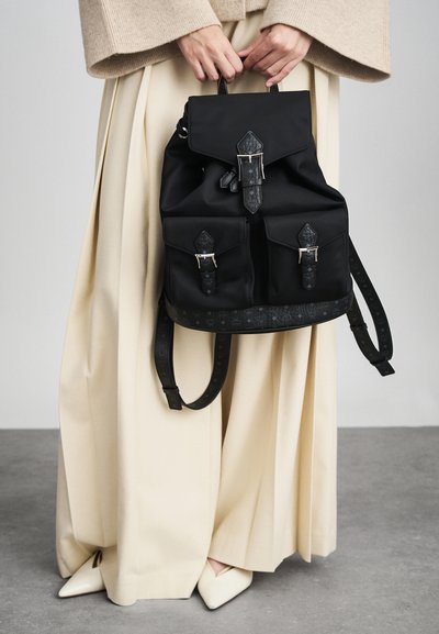 Persona con pantalones de pierna ancha beige y suéter sostiene una mochila negra con bolsillos frontales y cierres de hebilla, de pie sobre un suelo gris.
