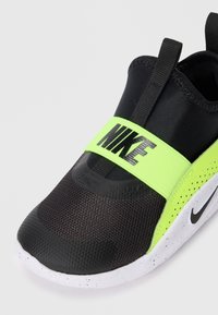 Chaussure de sport noire avec une tige texturée, une sangle vert fluo portant la marque "NIKE" et une semelle blanche mouchetée.