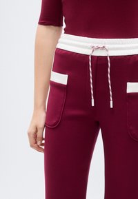 Μπορντό φόρμα με λευκή ελαστική μέση, κορδόνι και δύο εμπρόσθιες τσέπες με αντίθετη λευκή επένδυση. Ομαλή υφή υφάσματος.