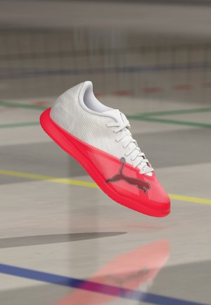 Λευκό και κόκκινο παπούτσι ποδοσφαίρου Puma για εσωτερικούς χώρους με μαύρο λογότυπο Puma να αιωρείται πάνω από το γυμναστήριο με έγχρωμες γραμμές γηπέδου.