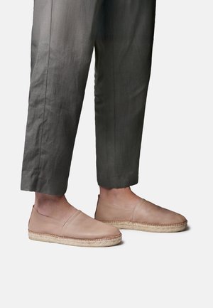 Nohy v krátkych tmavosivých nohaviciach a béžových kožených slip-on espadrilách s tkanou juta podrážkou na bielom pozadí.