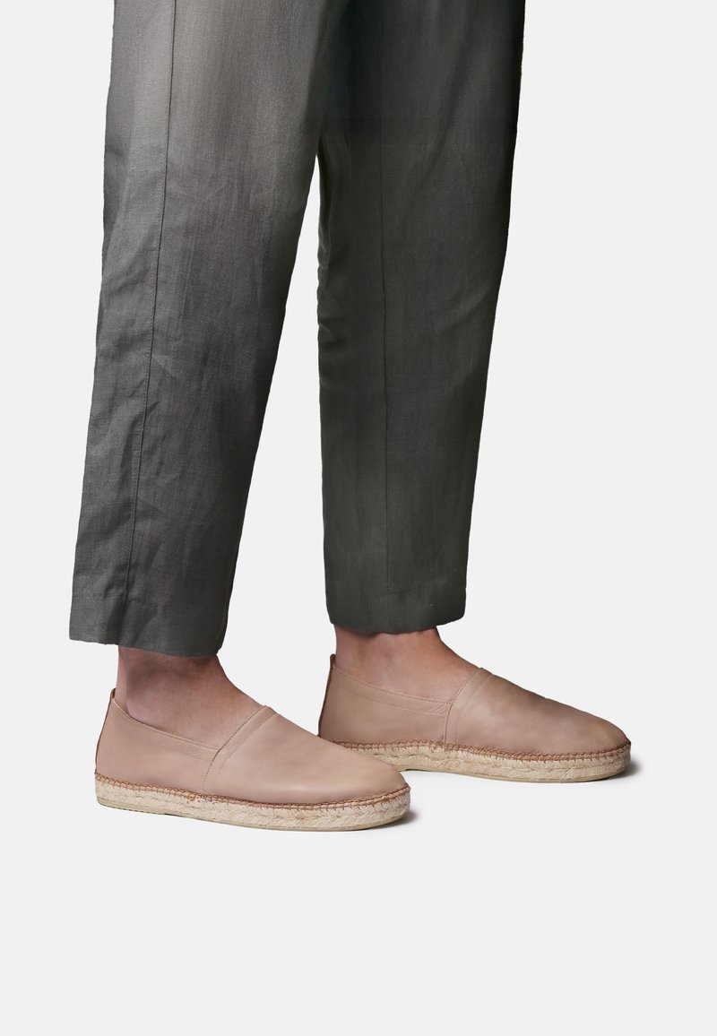 Jalad, mis kannavad lühendatud tumehalli pükse ja beeže nahast libisevaid espadrille koos kootud joodisäärega valgel taustal.