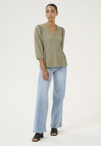 Blusa avvolgente verde oliva con maniche a palloncino di tre quarti, tessuto texturizzato, abbinata a jeans a gamba larga in lavaggio chiaro e sandali neri con tacco.