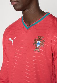Κόκκινη ποδοσφαιρική φανέλα με υφασμένο σχέδιο, λαιμόκοψη σε σχήμα V, τυρκουάζ γιακά, με το έμβλημα της εθνικής ομάδας της Πορτογαλίας και το λογότυπο της Puma.