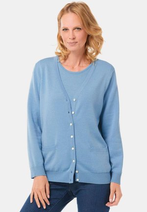 Hellblauer Cardigan mit V-Ausschnitt, Knopfschluss, zwei vorderen Taschen und gerippten Bündchen. Über einem passenden Hemd getragen.