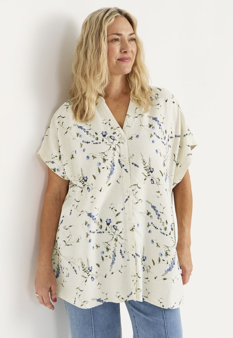 Kvinde med blondt, bølget hår iført en cremefarvet skjorte med blå blomsterprint og lyseblå jeans, stående op ad en hvid væg.