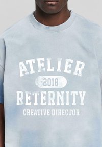 Light blue cotton t-shirt with faded white text: "ATELIER 2018 RETERNITY CREATIVE DIRECTOR." Short sleeves and crew neck design.
