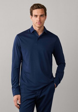 Jonge man met kort bruin haar, gekleed in een marineblauw poloshirt met lange mouwen en bijpassende broek, staand tegen een grijze achtergrond.