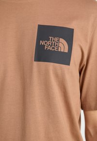 Camiseta de manga corta marrón con un diseño ajustado, que presenta un parche cuadrado negro en el pecho con el logo de "THE NORTH FACE" en dorado.