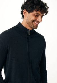 Svart höghalset zip-up tröja i stickad kvalitet. Har en slät yta och subtila detaljer på dragkedjans dragare.