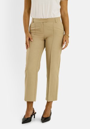 Mujer que lleva pantalones cortos de talle alto color beige con costuras frontales, camisa blanca metida, tacones negros de punta y pulseras y anillos dorados.