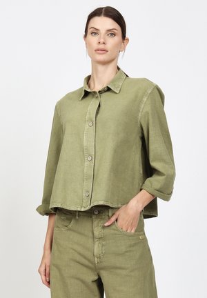 Woman with dark hair wearing a loose olive green button-up shirt with rolled sleeves and matching high-waisted pants, hand in pocket.
