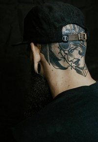 Gorra negra con textura de pana y hebilla de metal. En la parte posterior presenta un tatuaje detallado de la cara de una mujer con líneas marcadas y sombras.