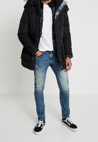 Giacca a vento nera con cappuccio foderato in pelliccia, indossata sopra una camicia bianca. Jeans chiari strappati e sneaker nere con lacci bianchi.