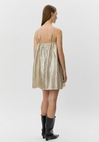 Glittrig guldig ärmlös klänning med en rynkad baksidesdesign, tillverkad av ett glänsande, reflekterande material, matchad med svarta knähöga stövlar.