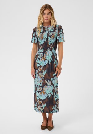Kvinde med blondt hår iført en kortærmet blå og brun blomsterprintet midi-kjole og brune spidse sko, stående mod en ensfarvet baggrund.