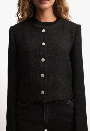 Femme portant une veste noire texturée avec six boutons décoratifs dorés sur le devant et des manches longues, associée à un pantalon noir.