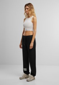 Weißes, kurzes Tanktop und schwarze Sweatpants mit Kordelzug. Die Hose verfügt über ein Logo-Patch. Die Schuhe sind beigefarbene Sneakers mit strukturierten Details.