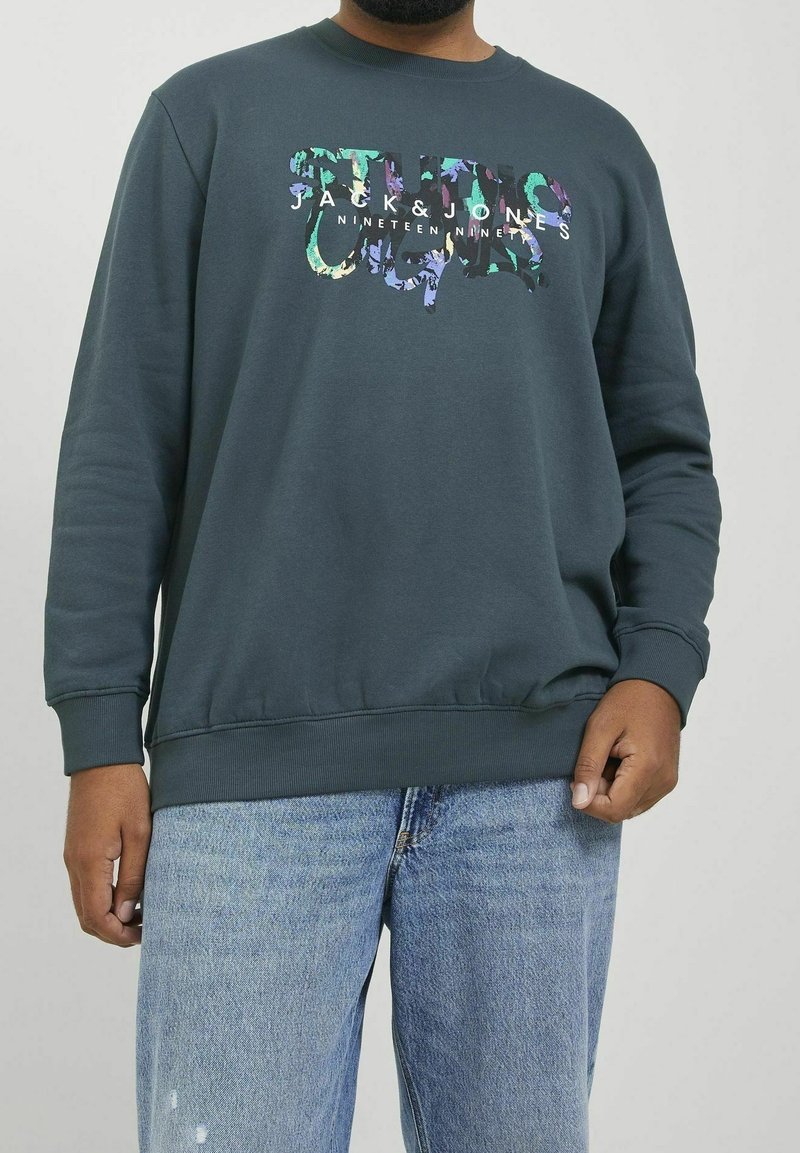 Homme portant un sweat-shirt vert foncé Jack & Jones avec un logo coloré et un jean bleu clair, debout devant un fond uni.