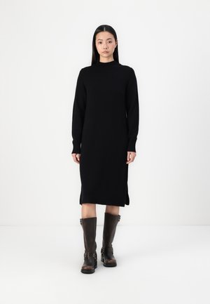 Black knitted dress with a high neck, long sleeves, and side slits; paired with knee-high brown leather boots featuring buckles.