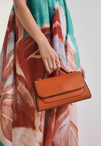 Orange läderhandväska med en vikt flik, prydd med svart kant och ett kort handtag, hålls mot en färgglad, mönstrad klänning.