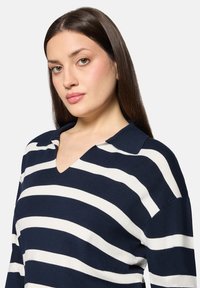 Pull-over à rayures bleu marine et blanches avec un col en V et des manches courtes. Confectionné en maille douce, il présente une coupe décontractée et un détail de col.