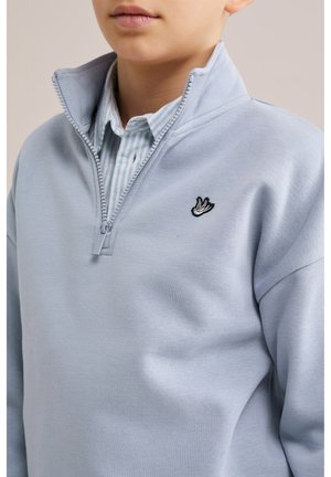 Garçon portant un sweat-shirt à fermeture éclair quart bleu clair sur une chemise à col rayée bleu et blanc, avec un petit logo d'oiseau noir et blanc sur la poitrine.