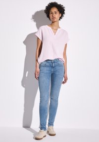 Haut rose clair à manches courtes avec un col en V, associé à un jean skinny bleu ajusté et des baskets beiges, sur un fond blanc uni.