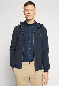 Marineblauwe hooded jack met rits, voorzakken en een subtiele logodetail. Draag het over een donker overhemd met knopen en beige broek.