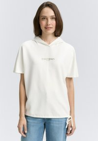 Vrouw in een witte hoodie met korte mouwen met de tekst "good times", staand tegen een effen lichte achtergrond, in een blauwe spijkerbroek.