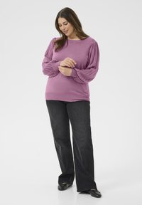 Kvinna som justerar ärmen på sin lila tröja med veckade ballongsärmar, iklädd svarta jeans och svarta lägenheter mot en neutral bakgrund.