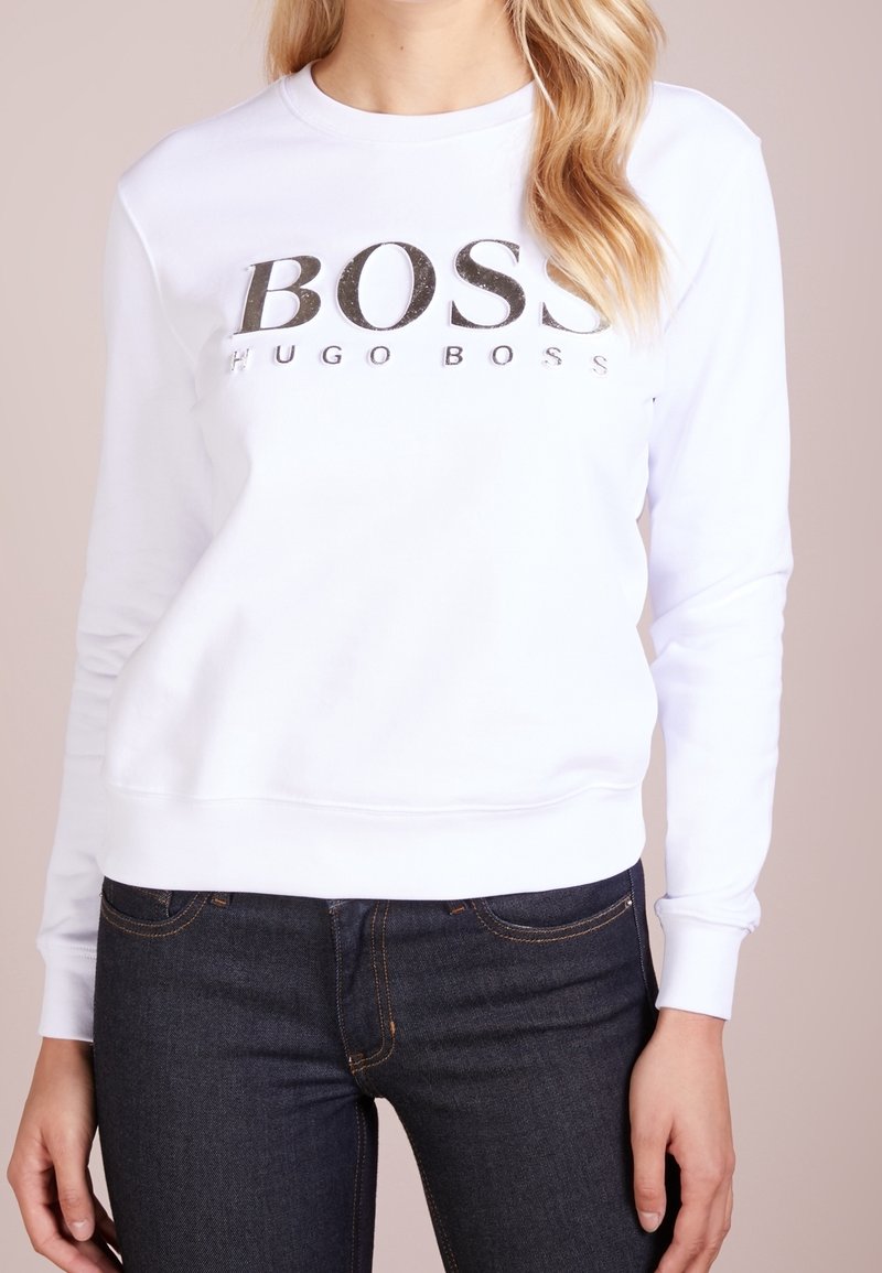 Sweat-shirt blanc à manches longues avec logo noir "BOSS HUGO BOSS", porté avec un jean bleu foncé sur une personne à la peau claire et aux cheveux blonds.