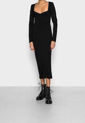 Schwarzes langärmliges Kleid mit quadratischem Ausschnitt und figurbetonter Silhouette, kombiniert mit schwarzen schnürbaren Kampfstiefeln, stehend auf einem hellen Boden.