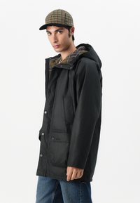 Μαύρο αδιάβροχο μπουφάν με κουκούλα, κούμπωμα με κουμπιά, τσέπη στο στήθος και καρό επένδυση. Συνδυάζεται με καπέλο με σχέδιο σε γήινες αποχρώσεις.